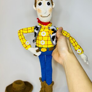 May include: Crocheted toy of Woody from Toy Story. He is wearing a yellow and red plaid shirt, a brown vest with a white star, blue jeans, and brown boots. He is holding his hat in his hand.