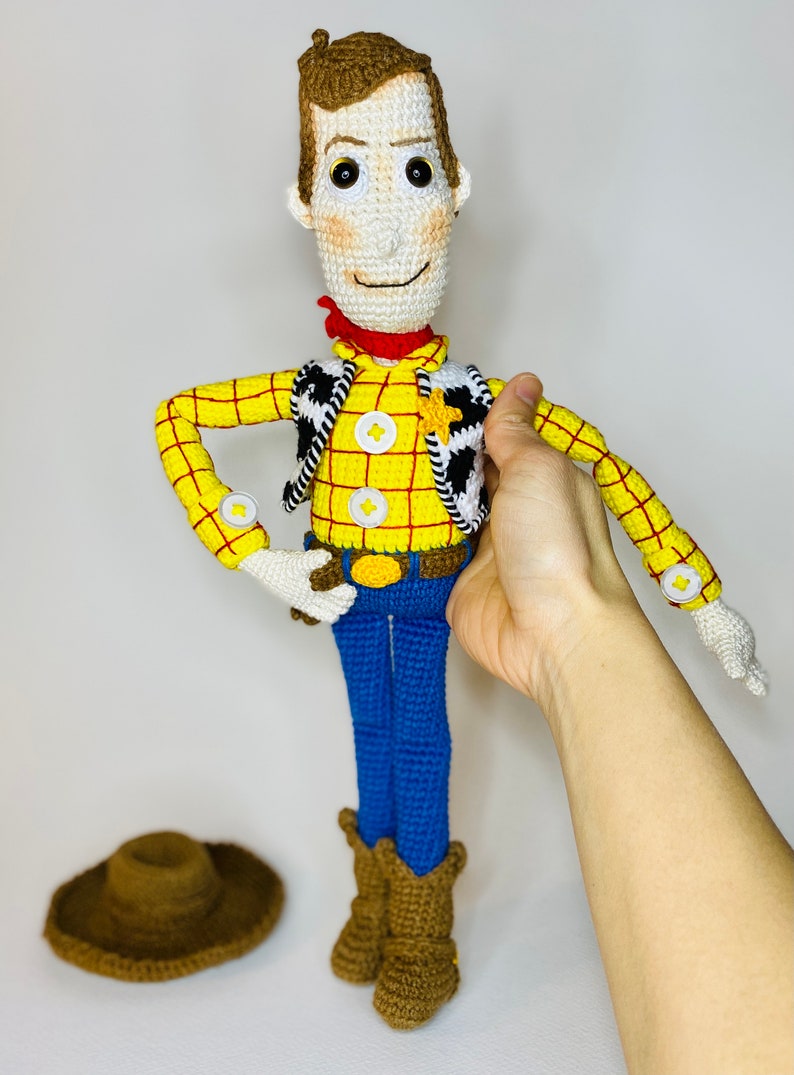 May include: Crocheted toy of Woody from Toy Story. He is wearing a yellow and red plaid shirt, a brown vest with a white star, blue jeans, and brown boots. He is holding his hat in his hand.