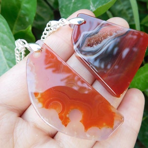 May include: Two agate stone pendants on silver chains. The pendants are both red and white with a natural agate pattern. One pendant is a half-circle shape and the other is a more irregular shape.