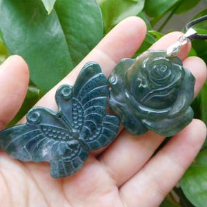 Puede incluir: Dos colgantes de piedras preciosas talladas. Uno es una mariposa con patrones de alas detallados, y el otro es una rosa. Ambos son de color verde oscuro con vetas verdes más claras. El colgante de rosa está sujeto a un cordón negro.