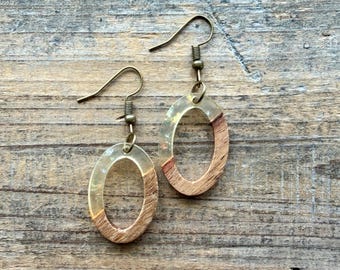 May include: A pair of oval-shaped dangle earrings featuring a combination of natural wood grain and translucent resin. Each earring is suspended from a bronze-toned metal hook, showcasing a smooth, polished finish on the lower wooden section.
