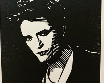 May include: Black and white woodcut print of a person's face. The person has dark hair and is looking down. The print is done in a simple style with bold lines.