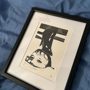 May include: Black and white print of a woman's face with a high heel shoe over her head. The print is framed in a black frame.