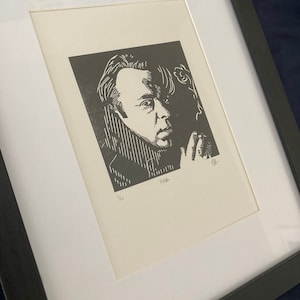 以下が含まれることがあります： 男性の肖像画の額装された白黒プリント。プリントは、白いマットが付いた黒いフレームに入っています。プリントには署名と1/10の番号が付けられています。男性はコートを着ており、真剣な表情をしています。