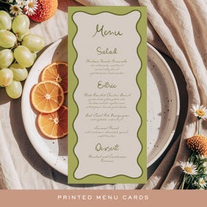 May include: A vertical menu card with a wavy green border, featuring menu items like salad and entree. The card is on a white plate with orange slices, grapes, and flowers. The text "PRINTED MENU CARDS" is at the bottom.