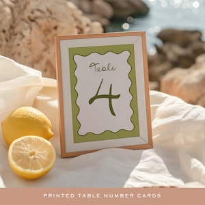 May include: A framed table number card with a light wood frame. The card has a white background with a green scalloped border and the number "4" in a dark green script. Two lemons are next to the card. The text "PRINTED TABLE NUMBER CARDS" is at the bottom.