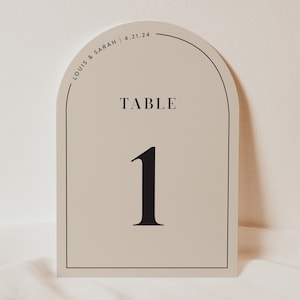 May include: Beige table number card with a black arch and the text "TABLE 1" in a black serif font. The top of the card has the text "LOUIS & SARAH | 4.21.24" in a black serif font.