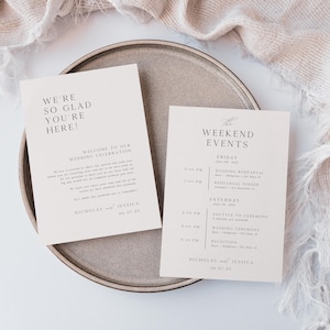 May include: Two cream-colored wedding cards with elegant typography, one reading "WE'RE SO GLAD YOU'RE HERE!" and the other "WEEKEND EVENTS." The cards are on a round, neutral-toned plate, with a textured fabric in the background.