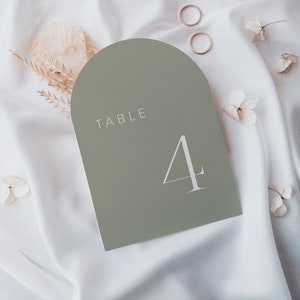 May include: A sage green table number card with rounded edges and a white arch at the top. The card says "TABLE 4" in white text.