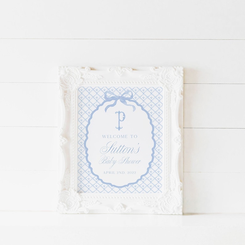 May include: A white frame with a blue and white patterned design. The design features a monogram "P" with a bow and the text "Welcome to Sutton's Baby Shower April 2nd, 2022".