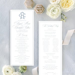 May include: Two rectangular wedding ceremony programs printed on white cardstock with light blue text and a monogram. The programs feature a detailed order of service and a list of the wedding party, accompanied by white ranunculus flowers.