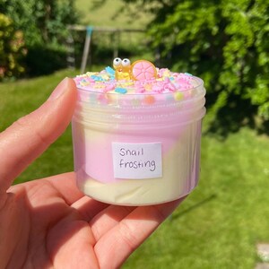 May include: A clear jar filled with a pink and white slime with colorful sprinkles on top. A small yellow snail with a pink shell is on top of the sprinkles. The jar has a label that says "Snail Frosting".
