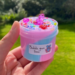 May include: A clear plastic container filled with pink and blue slime with a swirl design. The slime is topped with colorful sprinkles, gummy bears, and a label that reads "Bubble gum Bear".
