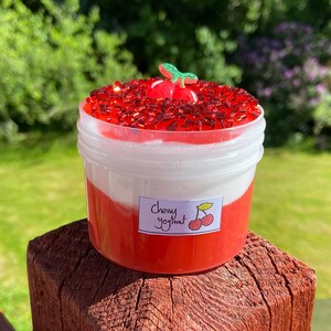 Puede incluir: Un recipiente de pl&aacute;stico transparente lleno de slime rojo y blanco, cubierto de purpurina roja y un amuleto de cereza verde. El recipiente tiene una etiqueta que dice "Cherry Yogurt".