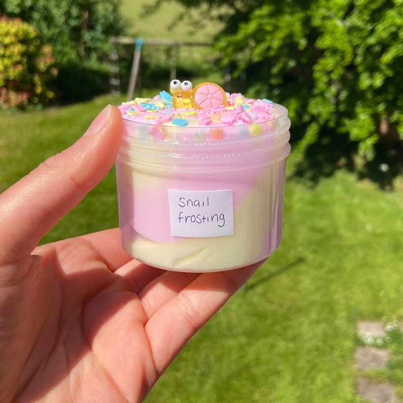 May include: A clear jar filled with a pink and white slime with colorful sprinkles on top. A small yellow snail with a pink shell is on top of the sprinkles. The jar has a label that says "Snail Frosting".