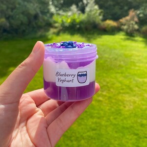 May include: A small clear jar with a purple lid filled with a layered purple and white slime. The jar has a label that says "Blueberry Yoghurt".