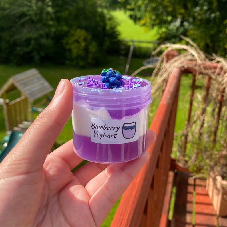 May include: A clear plastic jar filled with purple and white slime, topped with a cluster of blue blueberry-shaped charms and purple sprinkles. The jar has a white label that reads "Blueberry Yoghurt".