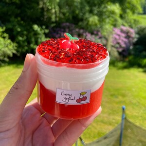 Puede incluir: Un recipiente de pl&aacute;stico transparente lleno de slime rojo y blanco. El slime est&aacute; cubierto con chispas rojas y una peque&ntilde;a cereza de pl&aacute;stico. El recipiente tiene una etiqueta que dice "Yogurt de cereza".