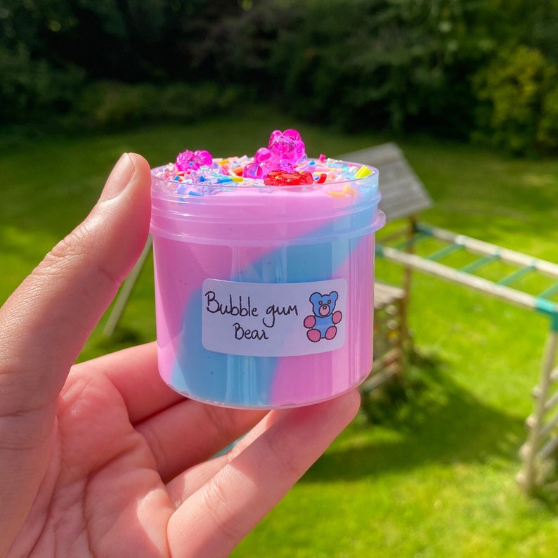 May include: A clear plastic jar filled with pink and blue slime with a swirl design. The slime is topped with colorful sprinkles and pink gummy bears. The jar has a white label that says "Bubble gum Bear".