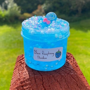 Puede incluir: Un tarro transparente lleno de slime azul con purpurina blanca e iridiscente. El slime tiene una etiqueta que dice "Blue Raspberry Slush" con un gr&aacute;fico de frambuesa azul.