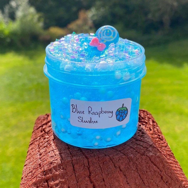 Puede incluir: Un tarro transparente lleno de slime azul con purpurina blanca e iridiscente. El slime tiene una etiqueta que dice "Blue Raspberry Slush" con un gr&aacute;fico de frambuesa azul.