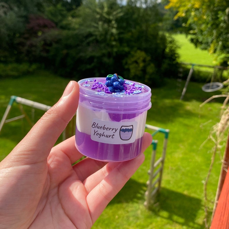 May include: A small clear jar filled with purple and white slime, topped with a cluster of blue blueberry-shaped charms and blue and white sprinkles. The jar has a label that reads "Blueberry Yoghurt".