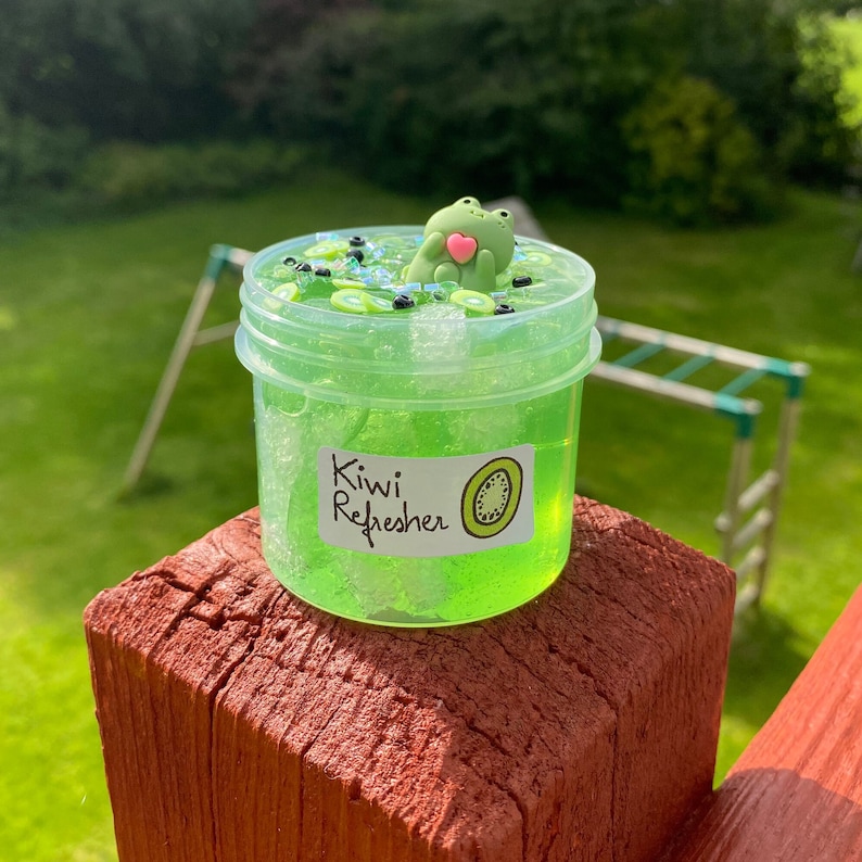 Pode incluir: Um frasco transparente de slime verde com glitter preto e prata, fatias de kiwi e um amuleto de sapo verde. O frasco tem um r&oacute;tulo branco que diz "Kiwi Refresher".