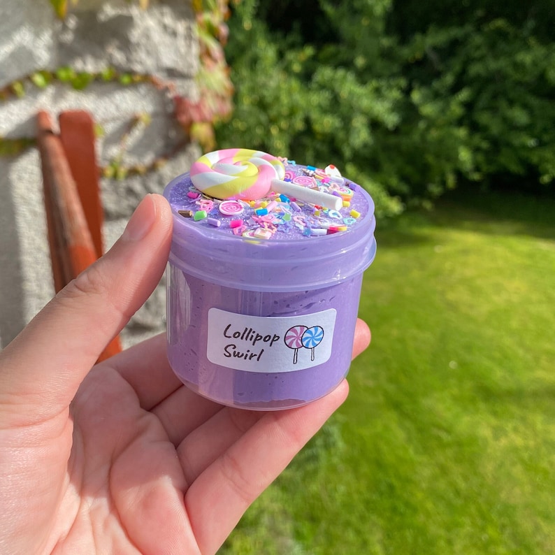 May include: Purple slime in a clear jar with a lollipop and sprinkles on top. The jar has a label that says "Lollipop Swirl".