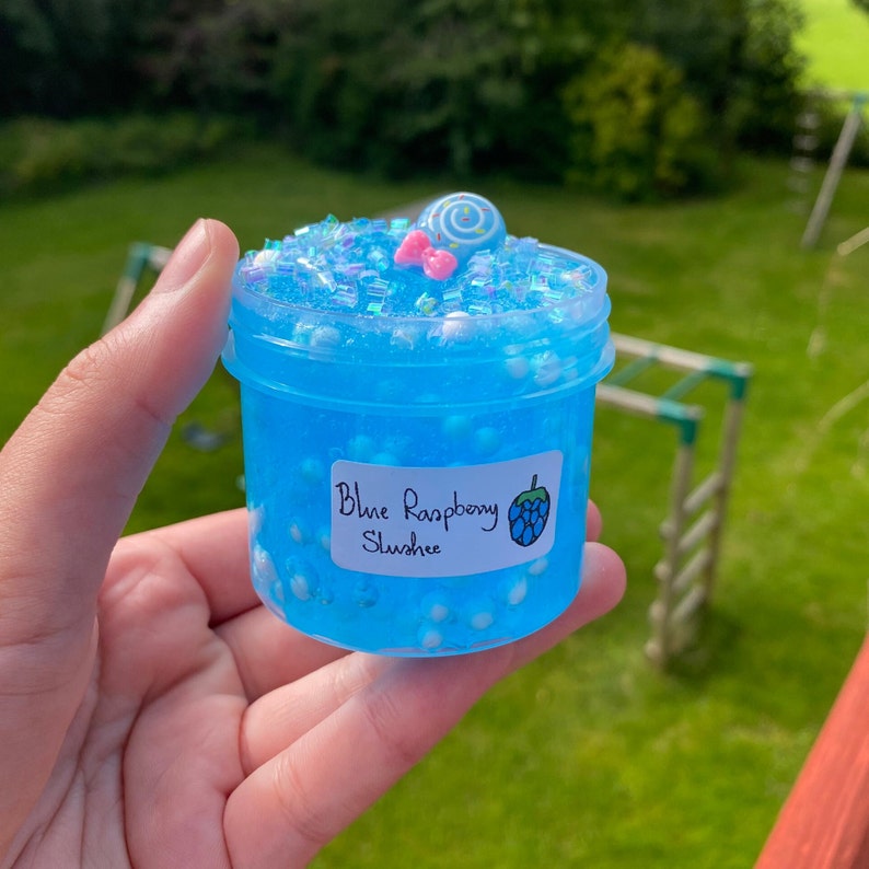 Puede incluir: Un peque&ntilde;o frasco transparente lleno de slime azul con purpurina azul y blanca y cuentas blancas. El frasco tiene una etiqueta blanca que dice "Blue Raspberry Slush" con un gr&aacute;fico de frambuesa azul.