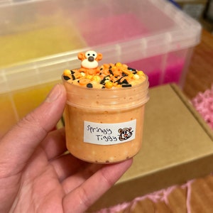 May include: Orange slime with black and gold sprinkles in a clear jar with a Tigger character on top. The jar has a label that says "Springy Tigger".