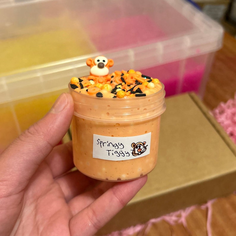 May include: Orange slime with black and gold sprinkles in a clear jar with a Tigger character on top. The jar has a label that says "Springy Tigger".