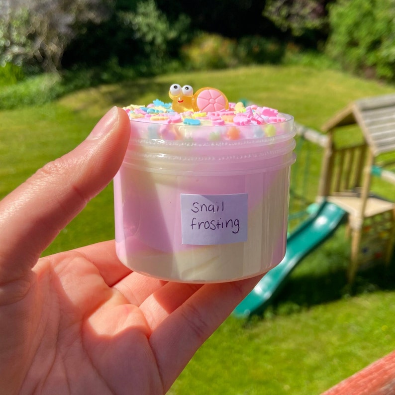May include: A clear plastic jar filled with pink and white slime with colorful sprinkles on top. A small yellow snail figurine with googly eyes sits on top of the sprinkles. The jar has a label that says "Snail Frosting".