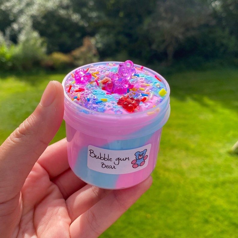 May include: A pink and blue slime with a swirl design in a clear plastic container. The slime is topped with colorful sprinkles and gummy bear charms. The container has a label that reads "Bubble gum Bear".