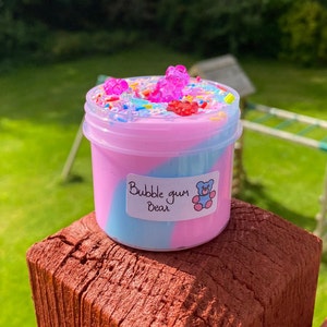 May include: A small clear jar filled with pink and blue slime with a swirl design. The slime is topped with colorful sprinkles and gummy bear shaped candies. The jar has a label that says "Bubble gum Bear".