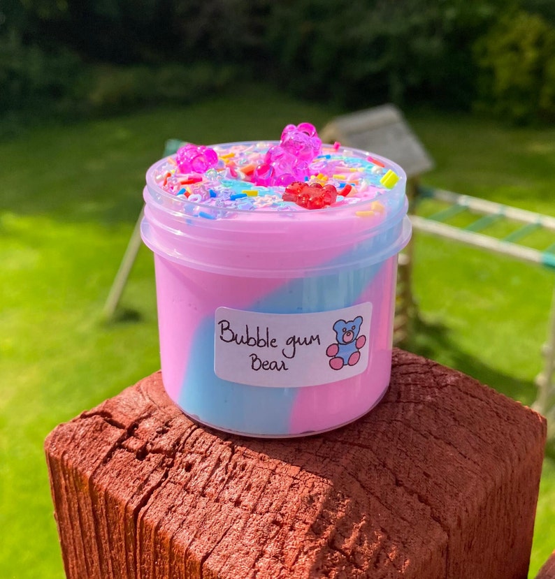 May include: A small clear jar filled with pink and blue slime with a swirl design. The slime is topped with colorful sprinkles and gummy bear shaped candies. The jar has a label that says "Bubble gum Bear".