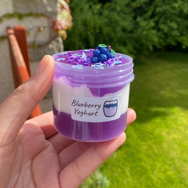 May include: A small clear jar with a purple lid filled with a white and purple layered slime. The slime is topped with blue and green blueberry-shaped decorations and purple and white sprinkles. The jar has a label that reads "Blueberry Yoghurt".