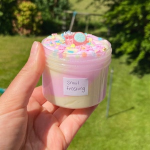 May include: A small clear jar filled with pink and white slime with colorful sprinkles on top. A small pink snail charm is on top of the sprinkles. The jar has a label that says "Snail Frosting".