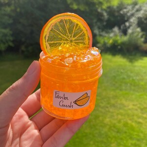 May include: Orange-flavoured slime in a clear jar with a glittery orange texture. The jar has a sticker that says "Fanta Crush" and a picture of orange slices.