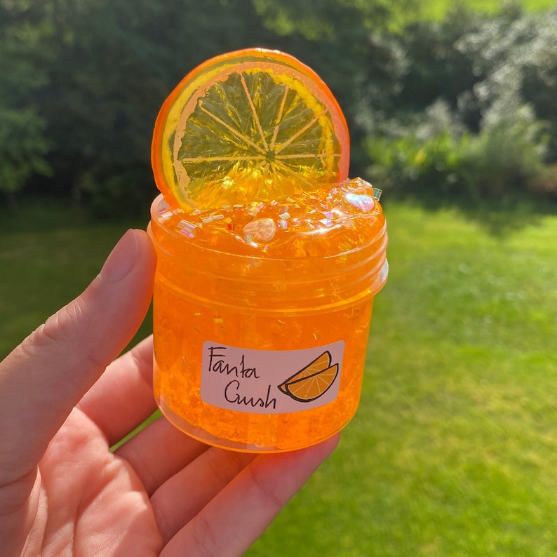 May include: Orange-flavoured slime in a clear jar with a glittery orange texture. The jar has a sticker that says "Fanta Crush" and a picture of orange slices.