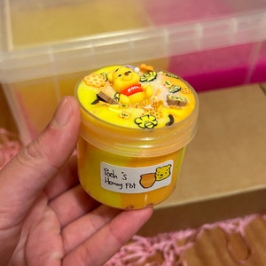 May include: A yellow and orange slime in a clear plastic jar with a Winnie the Pooh character on top. The jar has a label that says "Pooh's Honey Pot".