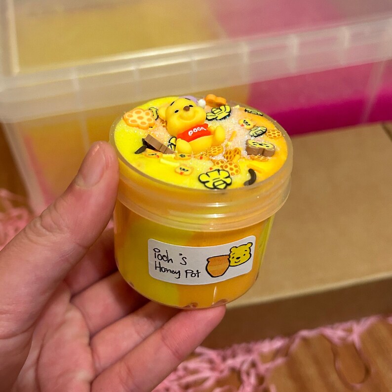 May include: A yellow and orange slime in a clear plastic jar with a Winnie the Pooh character on top. The jar has a label that says "Pooh's Honey Pot".