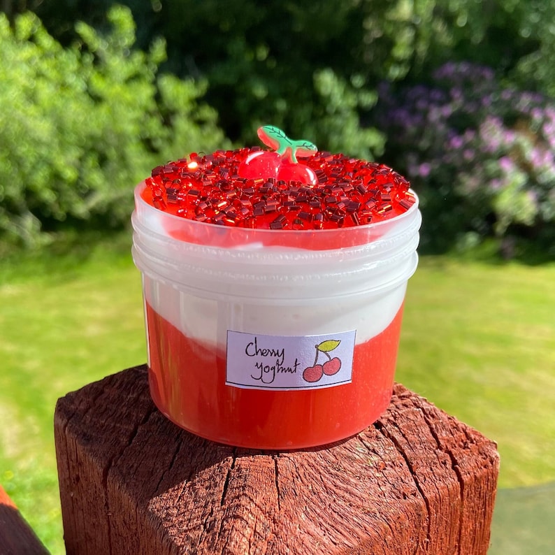 Puede incluir: Un recipiente de pl&aacute;stico transparente lleno de slime rojo y blanco, cubierto de purpurina roja y un amuleto de cereza verde. El recipiente tiene una etiqueta que dice "Cherry Yogurt".