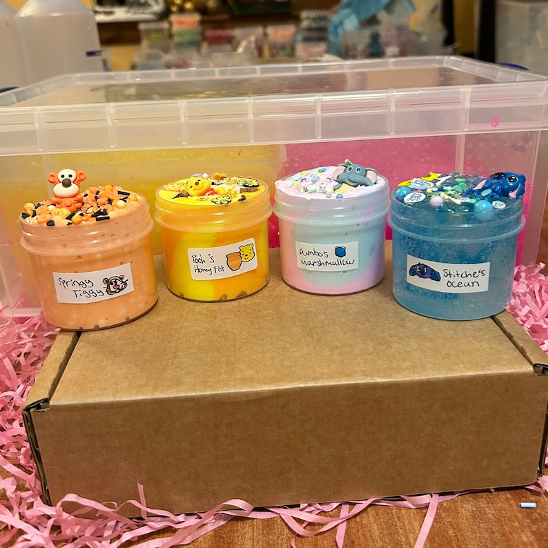 May include: Four small jars of slime, each with a different color and theme. The jars are labeled "Springy Tiggy", "Pooh's Honey Pot", "Rainbow Marshmallow", and "Stitch's Ocean".