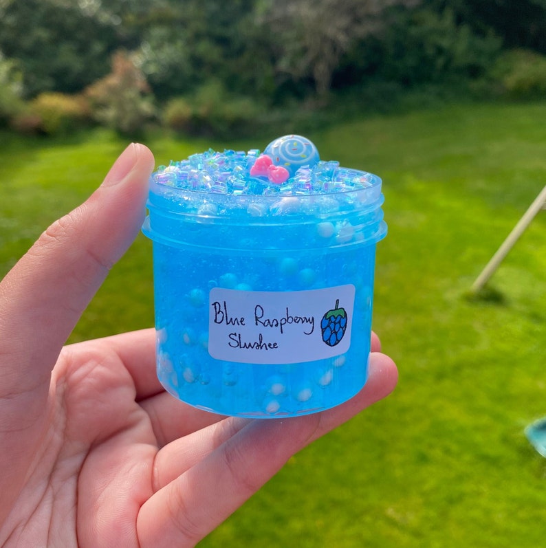 Puede incluir: Un tarro transparente lleno de slime azul con cuentas blancas y purpurina. El tarro tiene una etiqueta que dice "Blue Raspberry Slush" con un gr&aacute;fico de frambuesa azul.