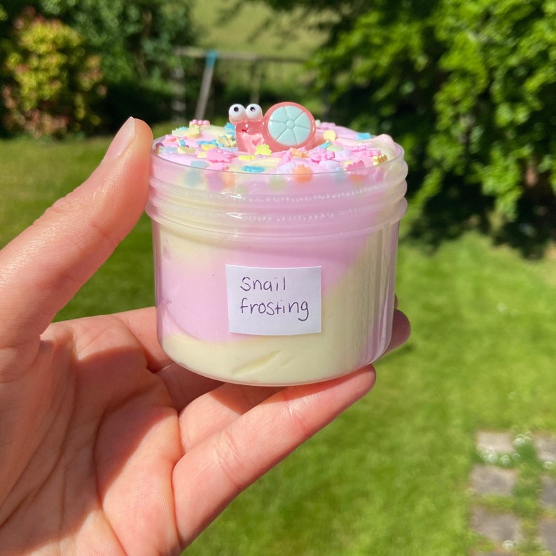 May include: A clear jar filled with pink and white slime with colorful sprinkles on top. A small plastic snail with googly eyes sits on top of the sprinkles. The jar has a label that says "Snail Frosting".