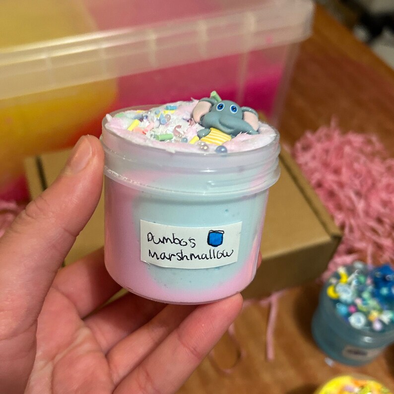 May include: A clear plastic jar filled with blue and pink slime with a small gray elephant figurine on top. The jar has a label that says "Dumbo's Marshmallow".