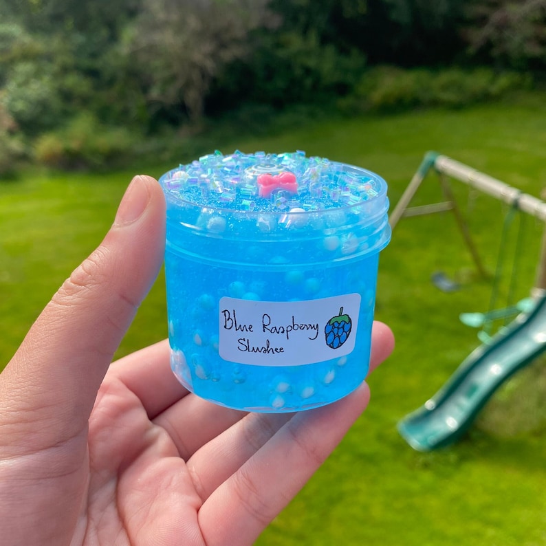 Puede incluir: Un peque&ntilde;o frasco transparente lleno de slime azul con purpurina blanca e iridiscente. El frasco tiene una etiqueta que dice "Blue Raspberry Slusher" con un gr&aacute;fico de frambuesa azul.