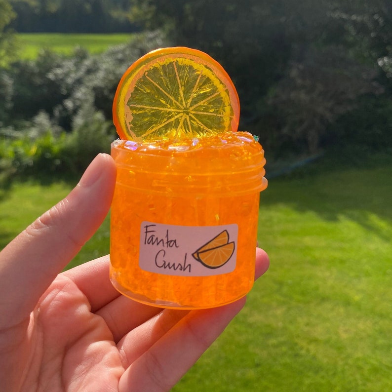 May include: Orange-flavoured slime in a clear jar with a plastic orange slice on top. The jar has a label that says "Fanta Crush" with a drawing of an orange slice.