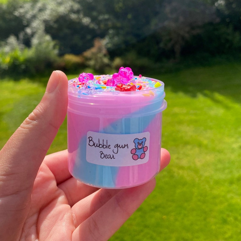 May include: A pink and blue slime with a gummy bear topping in a clear jar. The jar has a label that says "Bubble gum Bear".