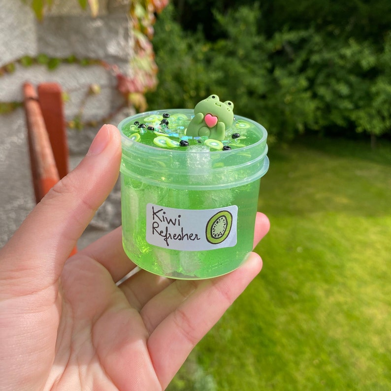 Pode incluir: Um frasco de pl&aacute;stico transparente cheio de slime verde, fatias de kiwi e contas pretas. Uma figura de sapo verde com um cora&ccedil;&atilde;o rosa flutua no topo do slime. O frasco tem um r&oacute;tulo branco que diz "Kiwi Refresher".